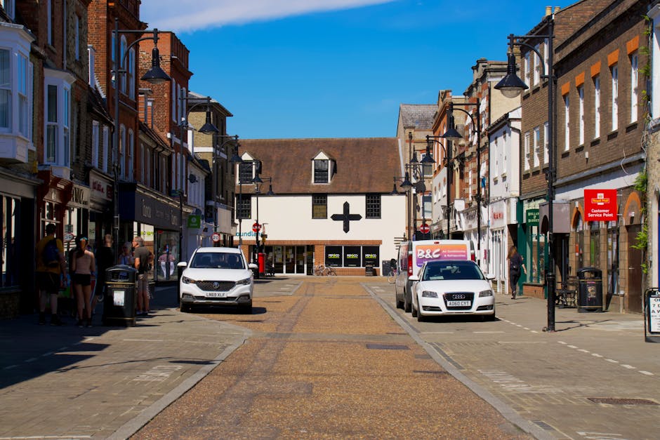 A daytime street scene on Acton High Street featuring a pedestrianized area with several shops lining both sides of the street and a central paved walkway. In the foreground, parked vehicles including a white van, an Audi car, and a boxed van are positioned along the curb on the right side, while public waste bins and streetlamps are visible. A group of people stand on the pavement near shop entrances on the left, some appearing to wait or chat. In the background, a small building with a large black cross on its white facade can be seen, suggesting a medical or pharmacy store. The sky is bright with a few clouds, and the overall scene represents a busy shopping area suitable for discussions regarding home relocation, furniture transport, or packing and moving services by companies like Acton Man and Van.