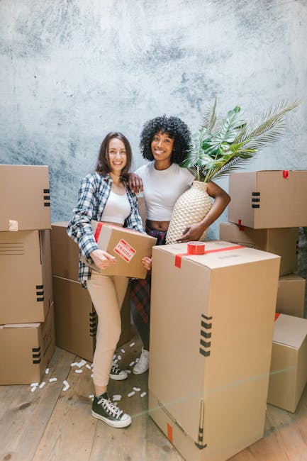 Two women are standing inside a property during a home relocation, surrounded by multiple cardboard boxes packed with household items. One woman is holding a medium-sized box with a red 'fragile' stic