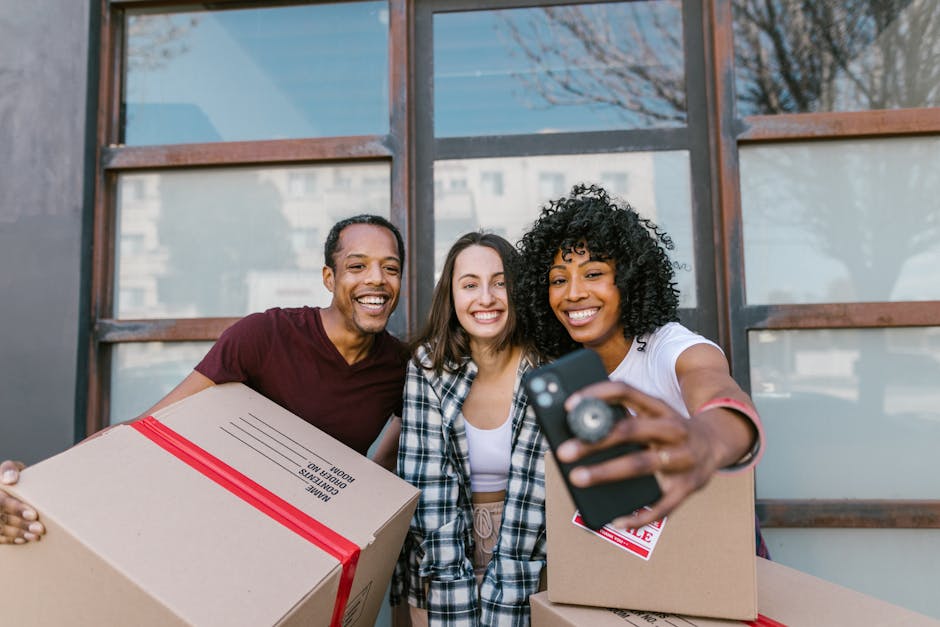 Two women are standing inside a property during a home relocation, surrounded by multiple cardboard boxes packed with household items. One woman is holding a medium-sized box with a red 'fragile' stic