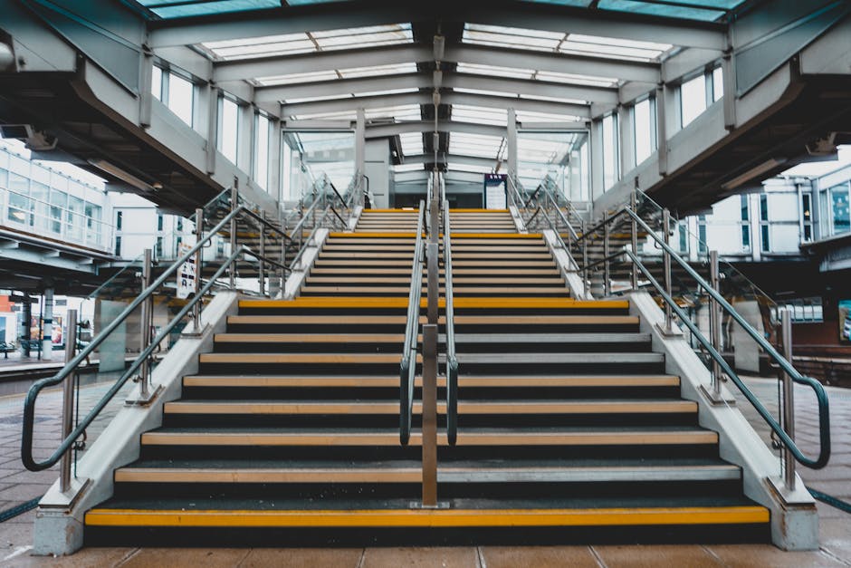 An exterior view of a set of outdoor stairs leading up to a modern, enclosed footbridge at Acton Town station, with metal handrails on both sides and yellow safety strips on each stair tread. The stairs are made of concrete and wood, with the covered structure overhead featuring glass panels allowing natural light to illuminate the area. In the background, portions of the surrounding station building and platforms are visible, with some signs and a few pedestrians near the station entrances. The scene depicts a typical station step-up that house removals companies like Acton Man and Van might use when planning access and logistics for home relocation or furniture transport, highlighting the importance of clear pathways for loading and unloading during moving services.