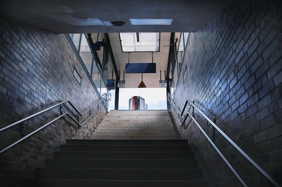The image shows a downward view of a staircase inside a building leading to the entrance or exit area, with brick walls on either side and metal handrails along both sides. The stairs are made of dark material, possibly tile or stone, with a small landing at the top where a glass door allows natural light to enter the space. Visible outside the glass door are signage panels and a partial view of the street or platform, indicating that this area is part of a transportation hub like a train or underground station. The lighting is primarily natural, coming from outside through the glass door, creating shadows on the stairs. This scene illustrates the interior access point used for home relocation or furniture transport processes, and it is relevant to the context of house removals or moving logistics, as handled by companies such as Acton Man and Van.