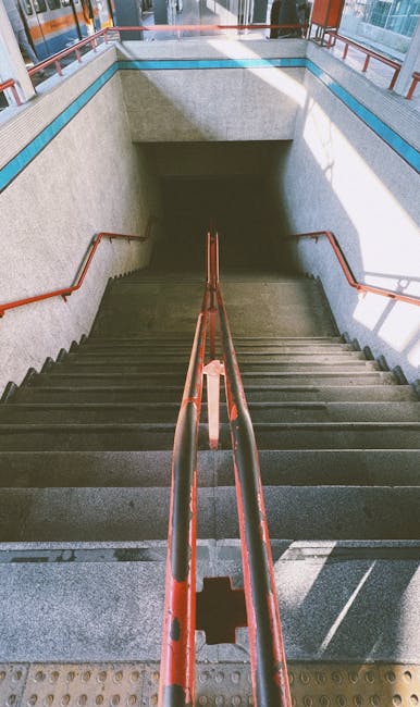 An exterior view of a set of outdoor stairs leading up to a modern, enclosed footbridge at Acton Town station, with metal handrails on both sides and yellow safety strips on each stair tread. The stairs are made of concrete and wood, with the covered structure overhead featuring glass panels allowing natural light to illuminate the area. In the background, portions of the surrounding station building and platforms are visible, with some signs and a few pedestrians near the station entrances. The scene depicts a typical station step-up that house removals companies like Acton Man and Van might use when planning access and logistics for home relocation or furniture transport, highlighting the importance of clear pathways for loading and unloading during moving services.